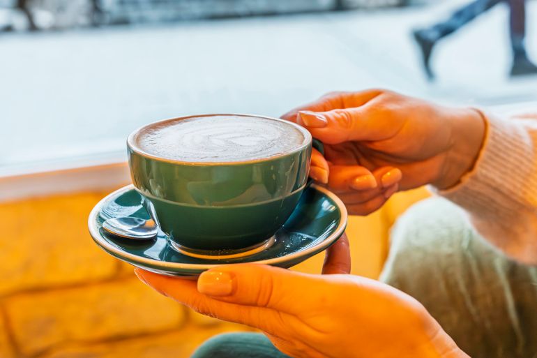 Young adult cradling warm green cup of latte with foam art, sitting in cozy cafe by sunlit window on a cold winter day, enjoying a peaceful, relaxing coffee moment